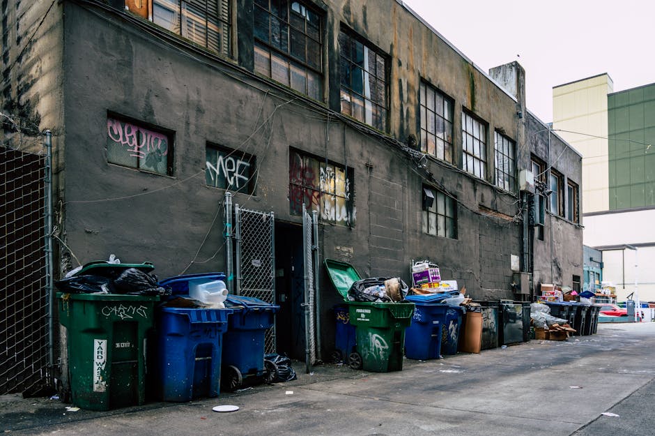 A large blue metal dumpster with visible rust and peeling paint, positioned on a paved surface in front of a modern building with dark horizontal siding and a section of blue tinted windows. The dumps