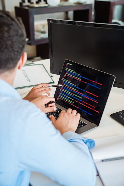 A person dressed in a light blue shirt is seated at a desk, working on a laptop displaying colorful lines of code on the screen. The individual is holding a pen in their right hand while using the key