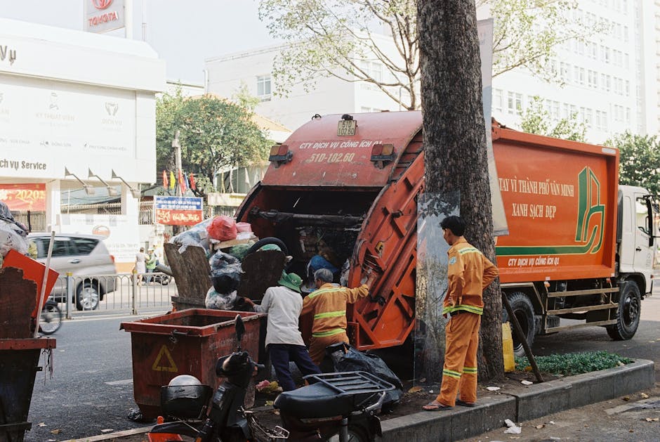 A waste collection worker wearing a black jacket and a high-visibility yellow vest with reflective strips is operating a rear-loading rubbish collection truck on a city street during dusk. The worker'