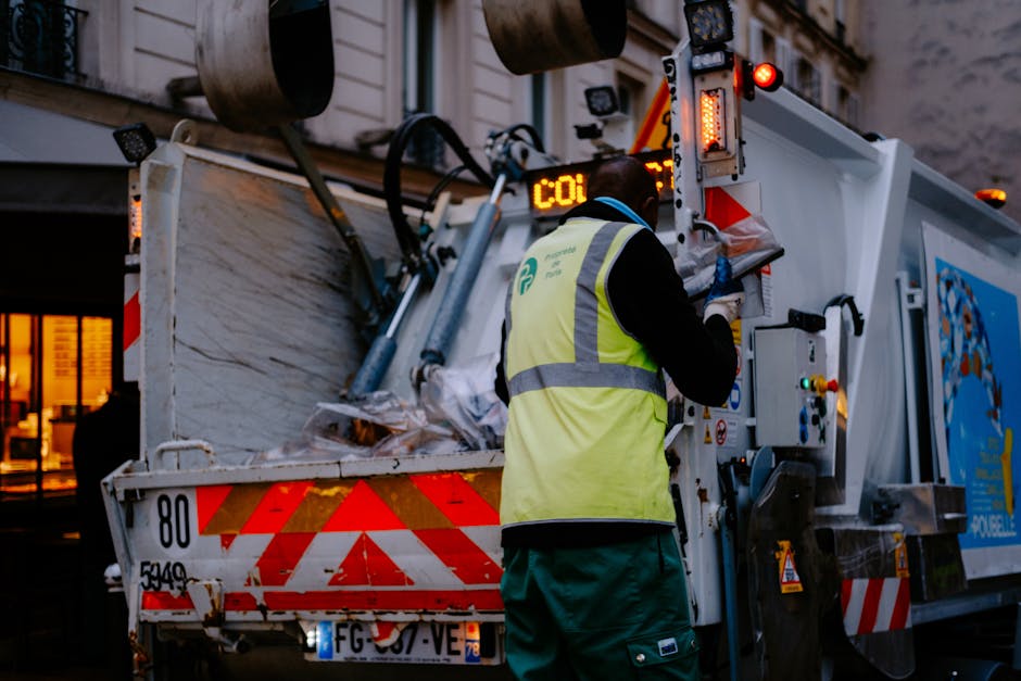 A waste collection worker wearing a high-visibility yellow vest and dark clothing operates controls on a commercial waste removal vehicle during evening hours, illuminated by the vehicle's lights. The