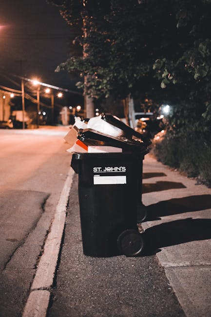 A waste collection worker wearing a high-visibility yellow vest and dark clothing operates controls on a commercial waste removal vehicle during evening hours, illuminated by the vehicle's lights. The