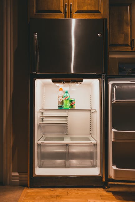 The image depicts a stainless steel, double-door refrigerator with a modern, sleek finish, positioned between wooden kitchen cabinets. The refrigerator door is open, revealing an empty interior with a
